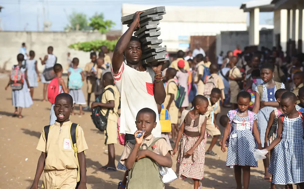 In Gonzagueville werden neue Klassenzimmer gebaut. | © UNICEF/DEJONGH Abidjan: Schulkinder und ein Mann, der die Bausteine für den Bau der Schule trägt
