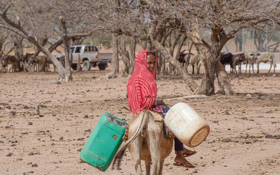 Ein Mädchen reitet auf dem Rücken eines Esels, mit sich trägt sie zwei Plastikkanister