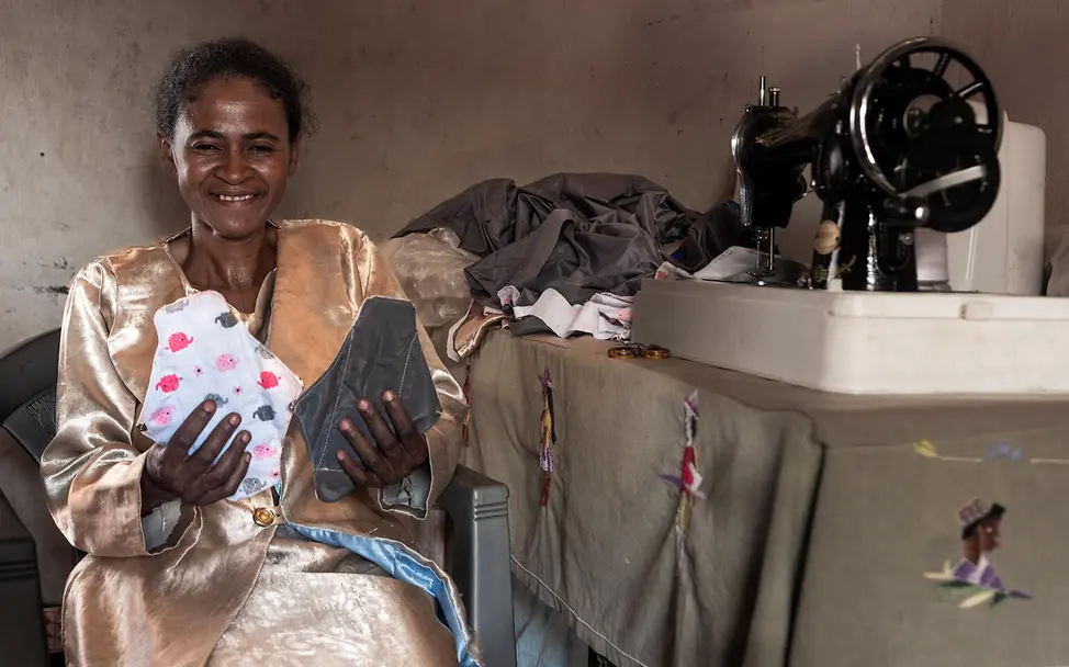 Schneiderin präsentiert ihre Arbeit | © UNICEF/UNI658459/Andrianantenaina Eine Schneiderin sitzt neben ihrer Nähmaschine und hält zwei Binden hoch, die sie genäht hat