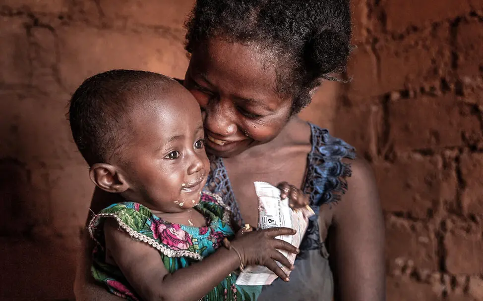 Mutter und Tochter in ihrem Zuhause | © UNICEF/UNI658451/Andrianantenaina Eine Mutter lächelt und drückt ihr Kleinkind an sich, welches therapeutische Zusatznahrung isst