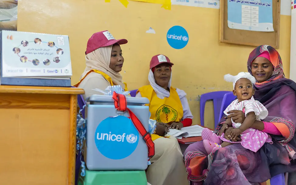 Eine Mutter sitzt mit ihrem Kind vor einer Kiste von UNICEF mit Impfstoff. 