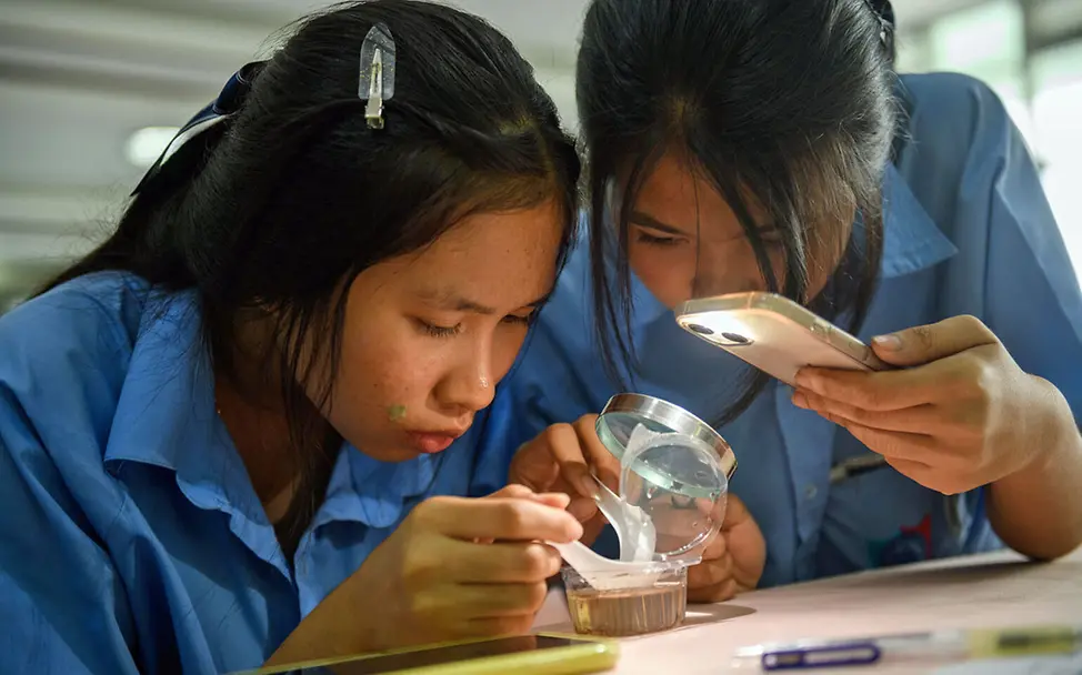 Malaria Kinder schützen: Zwei Schülerinnen in Thailand untersuchen mit einer Lupe und einer Handy-Taschenlampe Mückenlarven. 