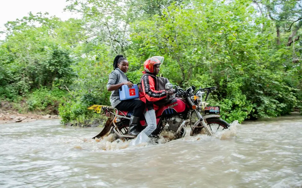 Eine Frau fährt mit einer Impfbox auf einem Motorrad durch einen überfluteten Fluss. I © UNICEF/Studio 19 Prisca ist Gesundheitshelferin im Nordwesten Tansanias und bringt Impfungen auch in die entlegensten Gebiete.