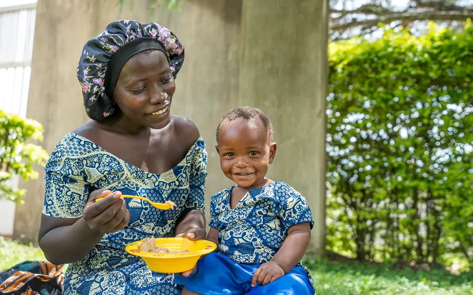 Uganda: Eine junge Mutter füttert ihr Kleinkind mit nährstoffreichem Brei. I © UNICEF/UNI786801/Abdul Eine Mutter füttert ihr einjähriges Kind in Uganda. Hilfsgüter wie Lebensmittel sind für kleine Kinder besonders wichtig.