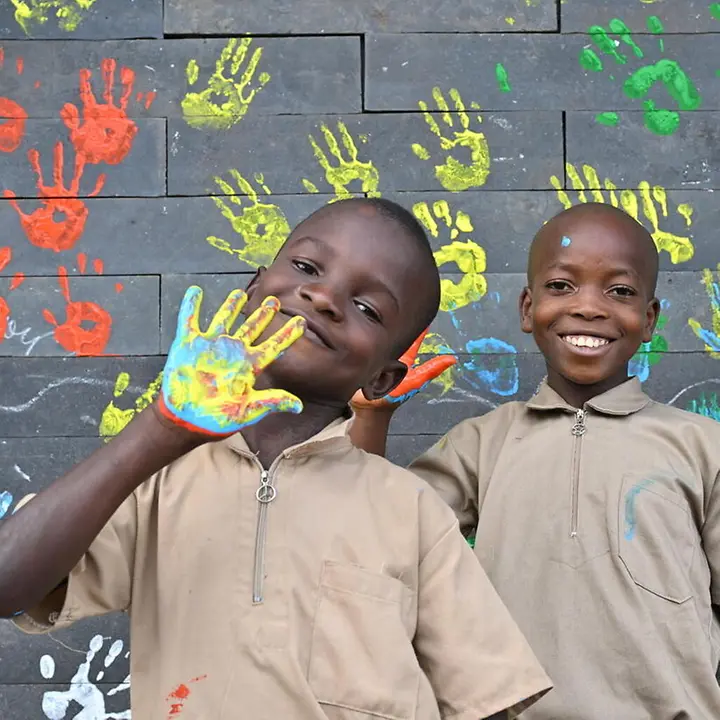 UNICEF baut in der Elfenbeinküste Schulen aus recycelten Plastikziegeln | © UNICEF/Dejongh Elfenbeinküste Bildung: Zwei Schulkinder zeigen ihre bemalten Handflächen vor einer Wand mit Handabdrücken.
