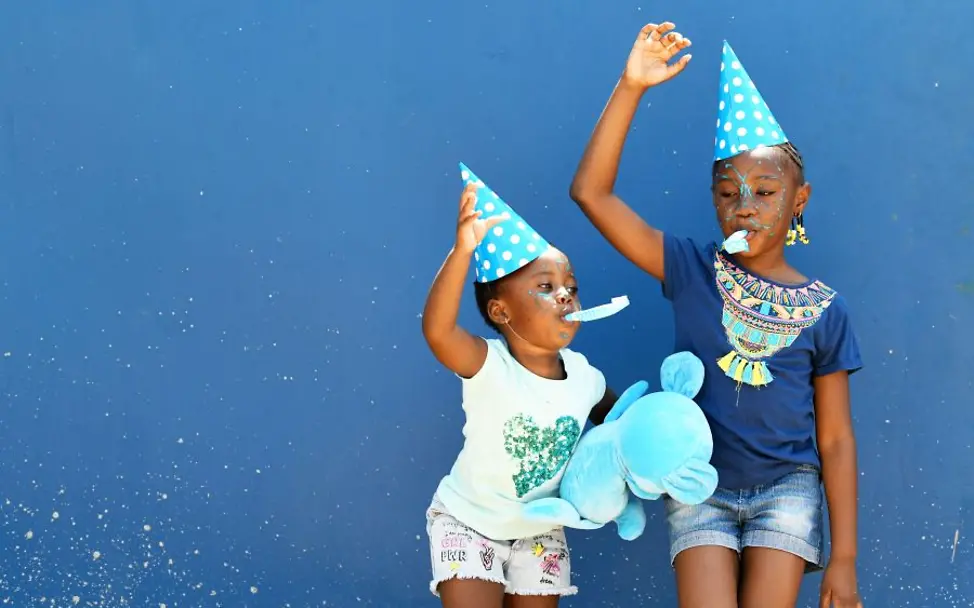 Kinderfreundliche Kommunen: Zwei Kinder feiern | © UNICEF/Dejongh Kinderfreundliche Kommunen: Zwei Kinder stehen vor einer blauen Wand, tragen Partyhüte und halten ein großes Plüschtier.