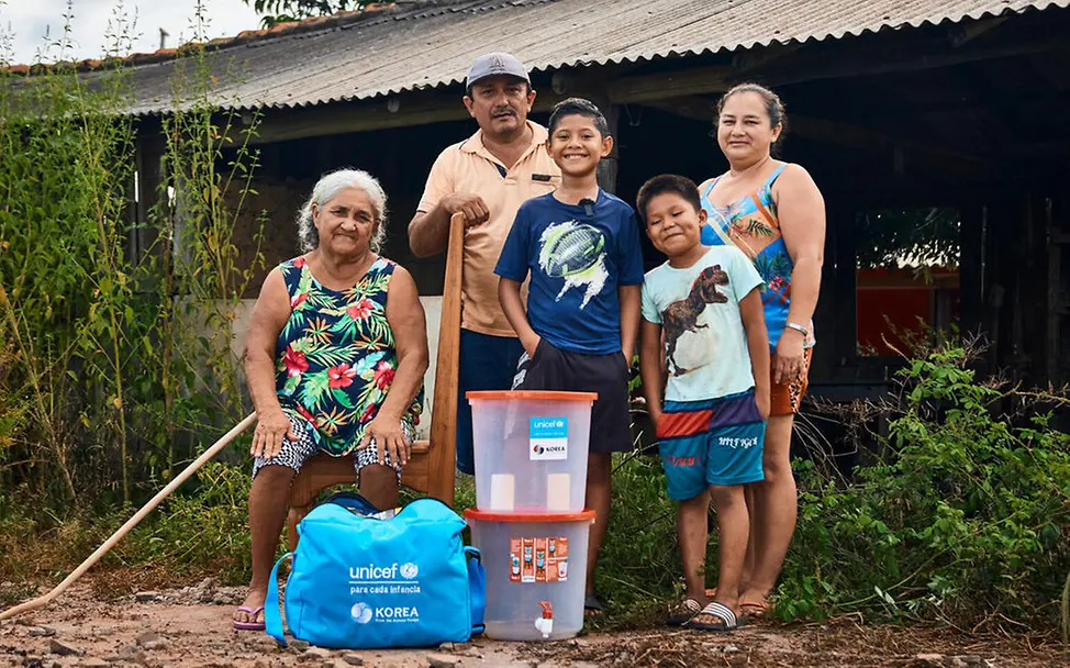 UNICEF versorgt Familien in Waldbrandgebieten mit Hygiene-Sets und anderen wichtigen Hilfsgütern. | © UNICEF/Soliz Waldbrand Folgen: Eine Familie steht mit UNICEF-Hilfsgütern vor ihrer Hütte.