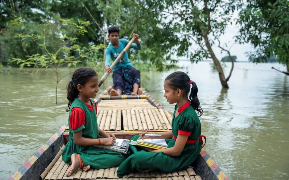 Durch Überflutungen wird vielen Kindern und Jugendlichen der Weg zu Bildungseinrichtungen abgeschnitten. | © UNICEF/Himu Bildung in Bangladesch: Zwei Mädchen sitzen mit Schulheften in einem Boot, das von einem Jungen gesteuert wird.