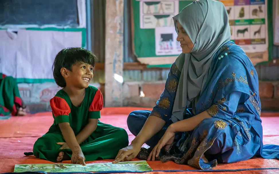 Let Us Learn UNICEF: Ein kleines Mädchen und eine Lehrerin sitzen auf dem Boden und schauen ein Buch an.
