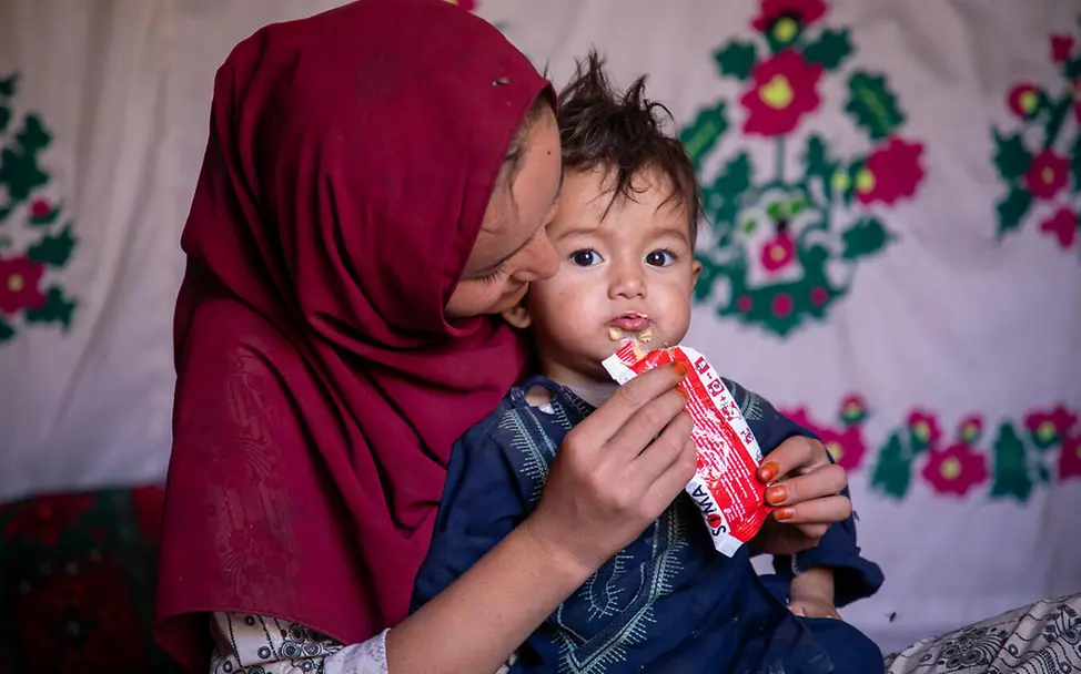 UNICEF liefert weltweit nahrhafte Erdnusspaste in Regionen, in denen Kinder hungern. | © UNICEF/Meerzad Was ist Hunger einfach erklärt: Ein Kind auf dem Schoß seiner Mutter wird mit Erdnusspaste gefüttert.