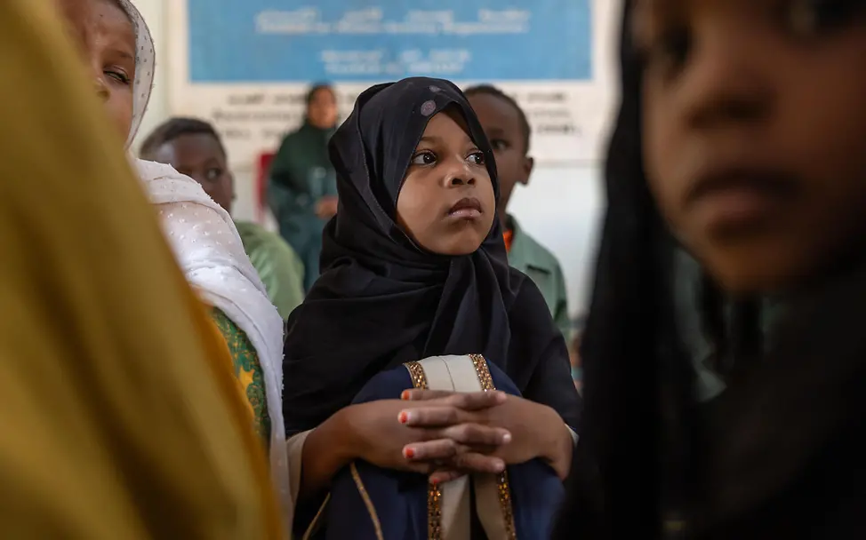 Sudan: Mädchen im Kopftuch sitzt und hört aufmerksam zu | © UNICEF/Elkhair Im Sudan sitzt ein Mädchen in dunklem Kopftuch und blauem Kleid mit goldenen Borten in einem Raum, neben anderen Kindern.